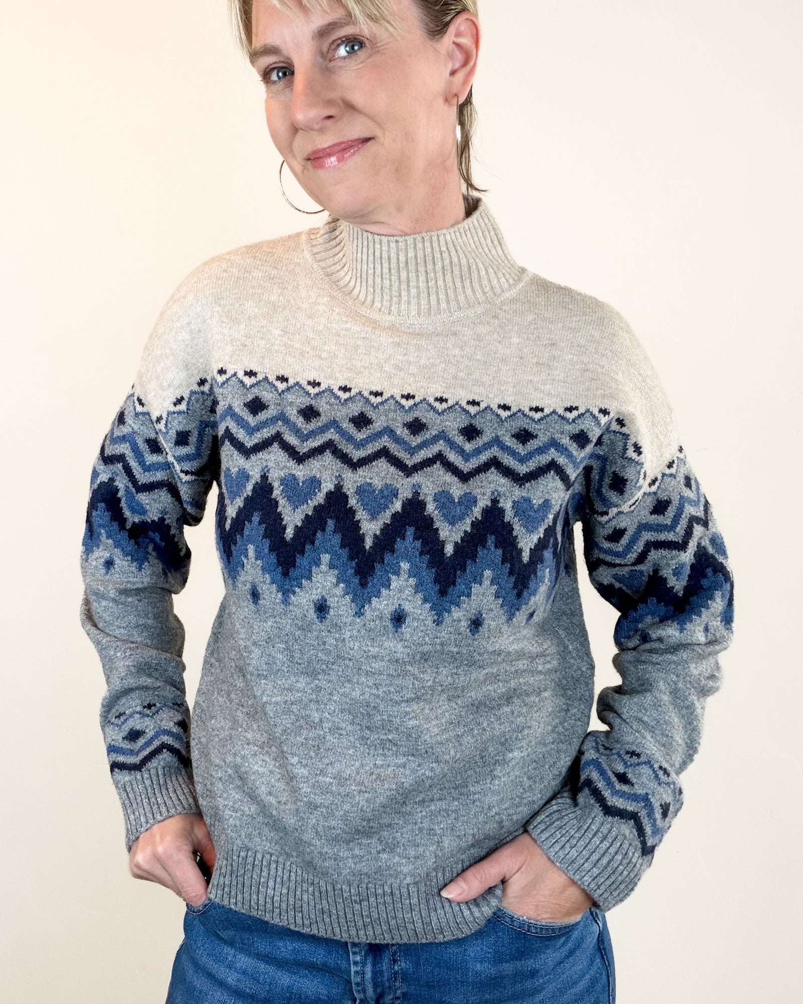 A woman wearing a gray and blue Fair Isle knit sweater with a turtleneck collar, paired with blue jeans, standing against a plain beige background.