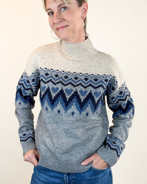 A woman wearing a gray and blue Fair Isle knit sweater with a turtleneck collar, paired with blue jeans, standing against a plain beige background.