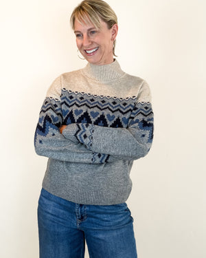 A woman wearing a gray and beige Fair Isle knit turtleneck sweater with blue patterned details, paired with blue jeans, standing against a plain off-white background.