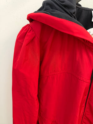 Close-up of a bright red hooded jacket with a black hood lining and black zipper detail, shown against a plain off-white background.
