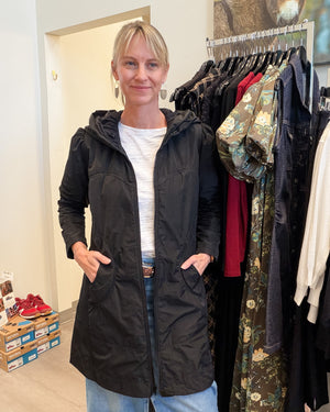 A woman wearing a black hooded jacket with front pockets, standing in a clothing store next to a rack of garments.