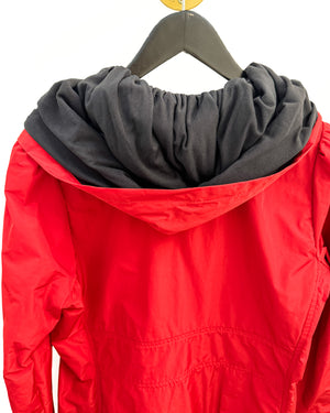 A close-up of a red hooded jacket with a gathered black hood, hanging on a black hanger against a plain white background.