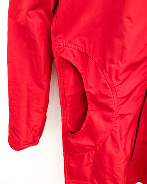 Close-up of a bright red lightweight jacket or coat showing the side seam, a large curved side pocket, and a black zipper detail against a plain white background.