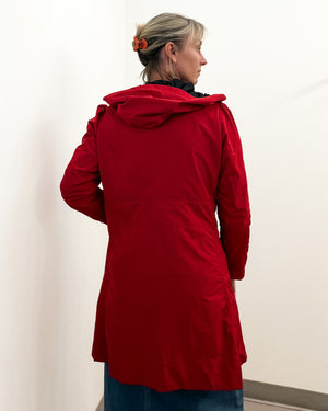 A woman wearing a bright red hooded jacket, shown from the back and side against a plain off-white wall.