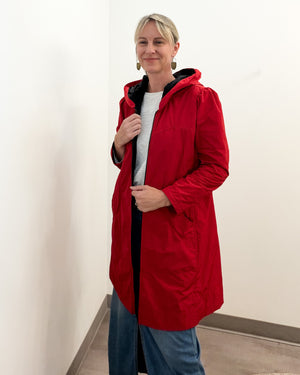 A woman wearing a red hooded jacket over a white top and blue jeans, standing in a plain indoor setting with off-white walls.