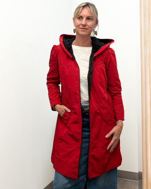 A woman wearing a bright red hooded jacket with a black inner lining, layered over a white top and blue jeans, standing against a plain white wall.
