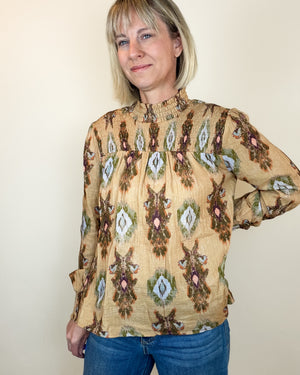 A woman wearing a beige long-sleeve blouse with a brown, green, and pink ikat-style geometric pattern, paired with blue jeans, standing against a plain beige wall.
