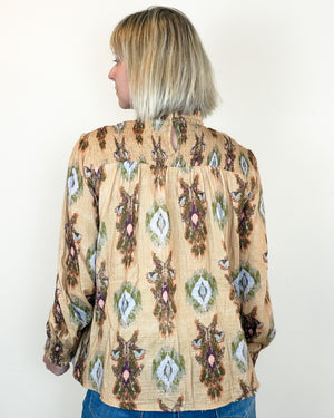 A woman viewed from the back wearing a beige long-sleeve blouse with a colorful geometric and floral pattern, paired with blue jeans against a plain white background.