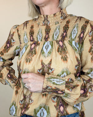 A close-up of a woman wearing a beige smocked blouse with a brown, green, and pink ikat-style pattern. The blouse features a high gathered neckline, a smocked chest panel, and long ruffled sleeves.