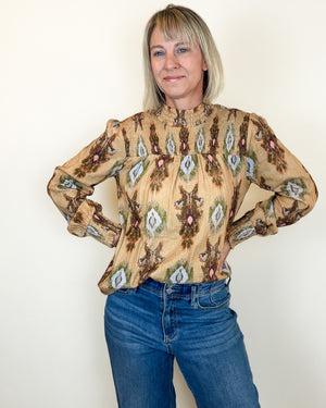 A woman with blonde hair wearing a beige long-sleeve blouse with a brown and green geometric pattern, paired with blue jeans, standing against a plain off-white wall.