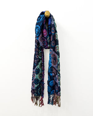 A long patterned scarf draped on a small wooden hanger against a plain white background. The scarf features a complex abstract design with swirling shapes in blue, purple, teal, and black, finished with fringed ends.