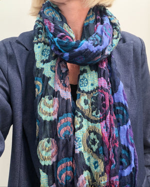 A close-up of a person wearing a dark scarf with a colorful paisley and medallion pattern in blue, purple, teal, pink, and beige, paired with a dark gray blazer.
