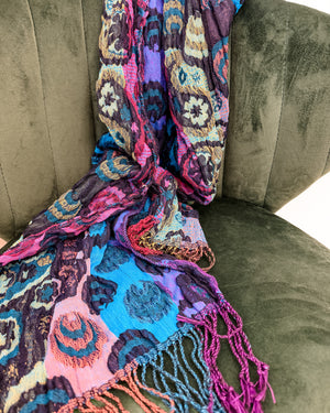 A multicolor patterned scarf with fringed edges draped over a dark olive green velvet armchair.