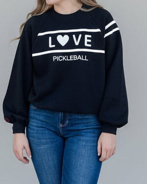 A woman modeling a black crewneck sweatshirt with 'LOVE PICKLEBALL' printed in white across the chest, paired with blue jeans and white flats, against a solid gray studio background.