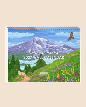 A spiral-bound 2027 National Parks wall calendar displayed against a solid beige background. The cover features a colorful landscape illustration of Mount Raina with green hills, wildflowers, pine trees, a wolf, and a bald eagle.