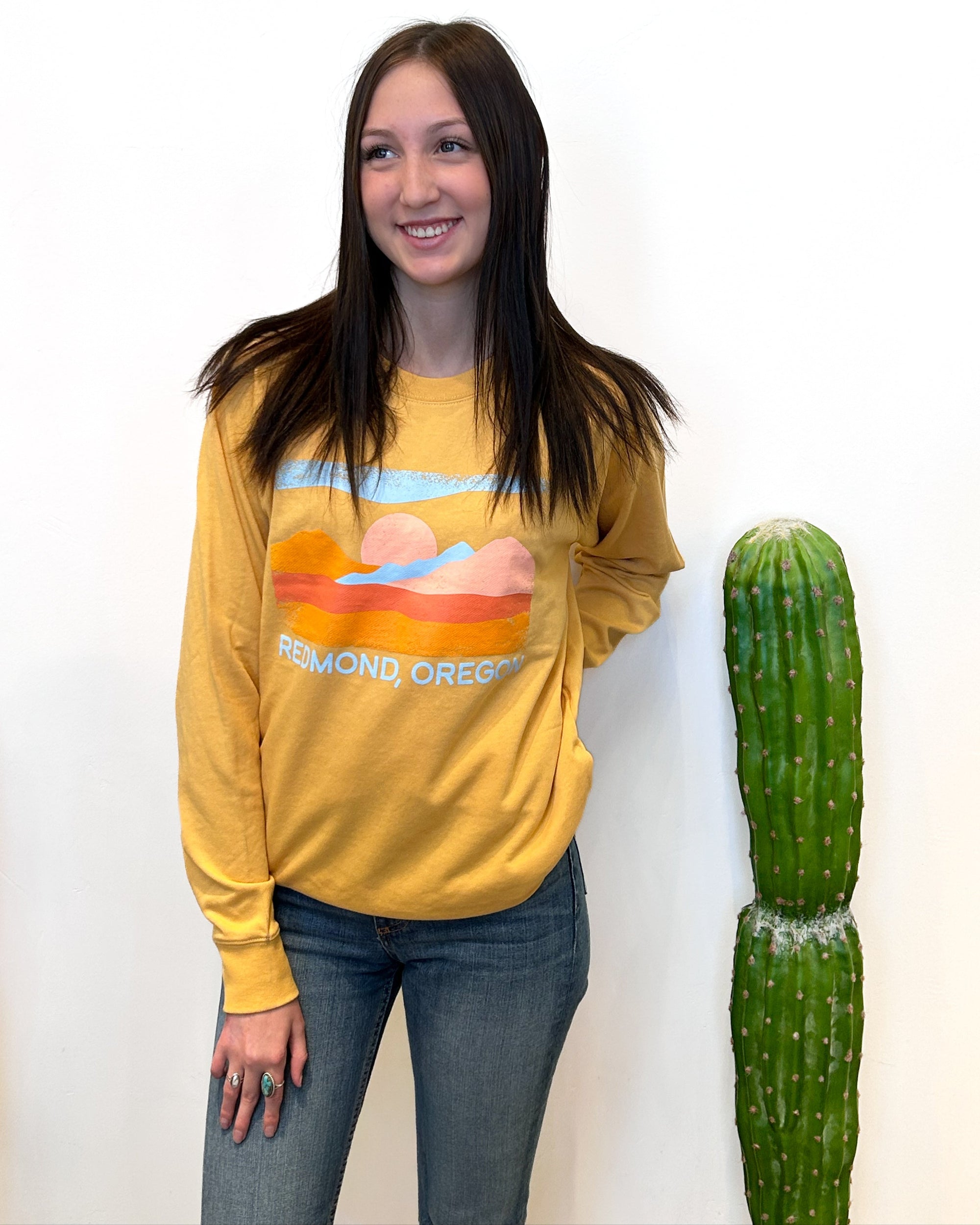 Person wearing a mustard yellow sweatshirt with a graphic design, standing next to a cactus on a white background