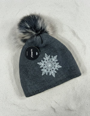 Dark gray knit beanie with a snowflake design and faux fur pom-pom on a wooden surface.