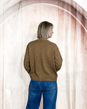 Person wearing a brown sweater and blue jeans standing against a plain background