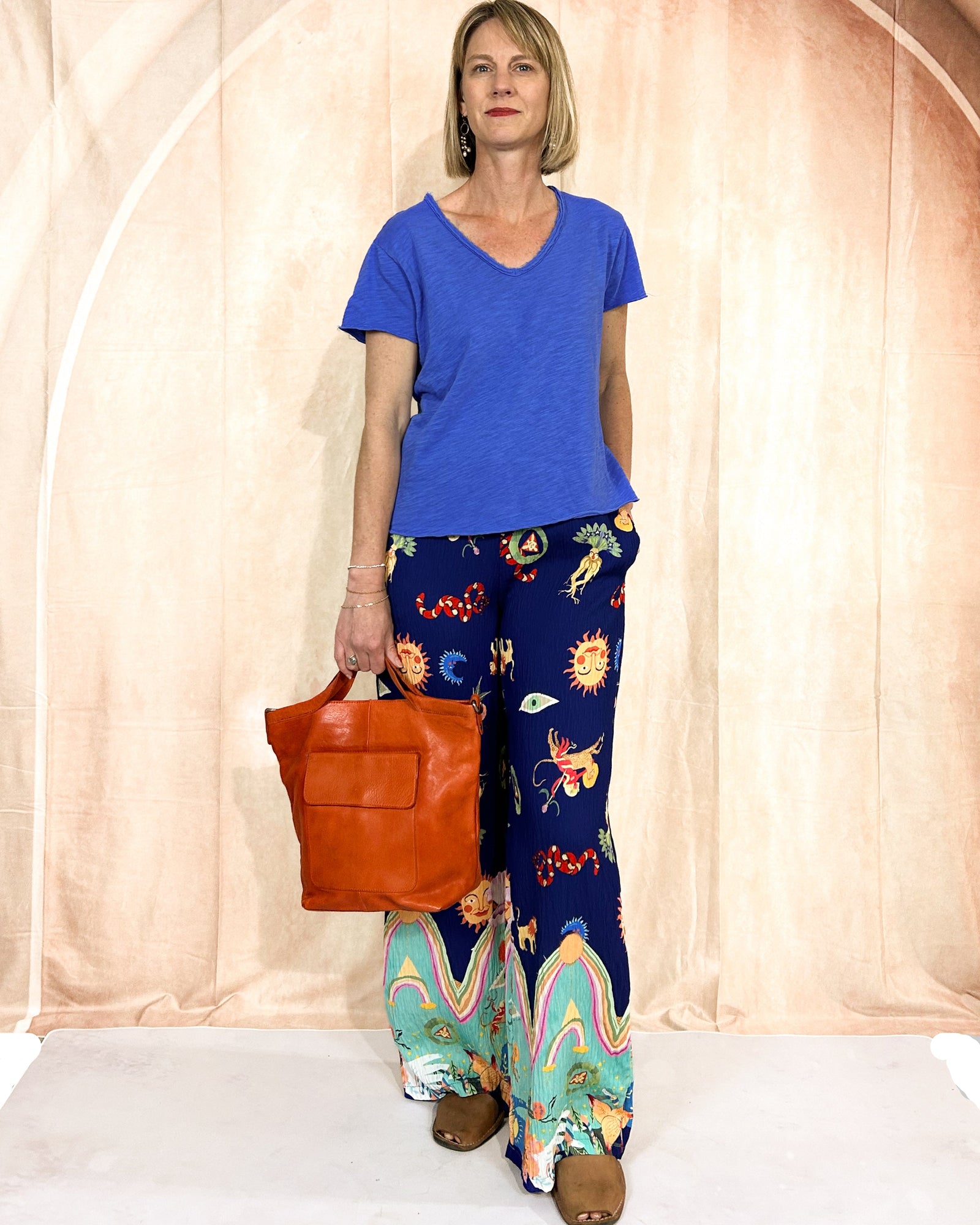Woman wearing a blue shirt and colorful pants, holding an orange bag against a plain background