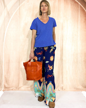 Woman wearing a blue shirt and colorful pants, holding an orange bag against a plain background