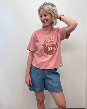 A woman modeling a pink crewneck t-shirt with a brown bear graphic and the word 'SMOKEY' on the front, paired with blue denim shorts, standing against a plain off-white wall.