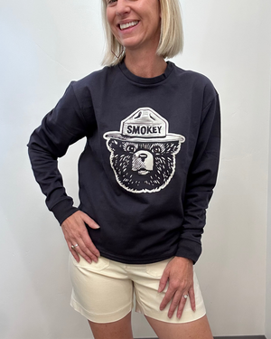 A woman wearing a navy long-sleeve sweatshirt with a white bear graphic and the word 'SMOKEY' on the front, paired with cream shorts, standing against a plain light gray wall.