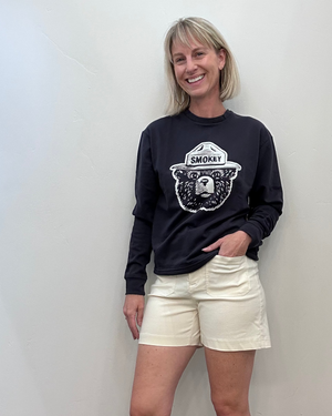 A woman stands against a plain off-white wall wearing a dark navy long-sleeve crewneck sweatshirt with a white graphic of a bear wearing a hat labeled 'SMOKY', paired with cream-colored shorts.