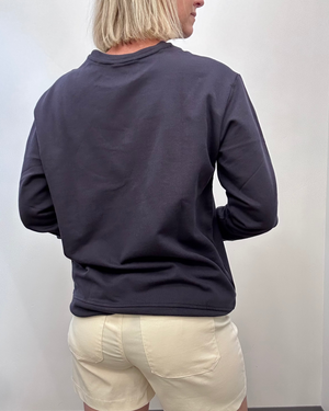 A person shown from the back wearing a navy crewneck sweatshirt and cream shorts against a plain light gray wall.