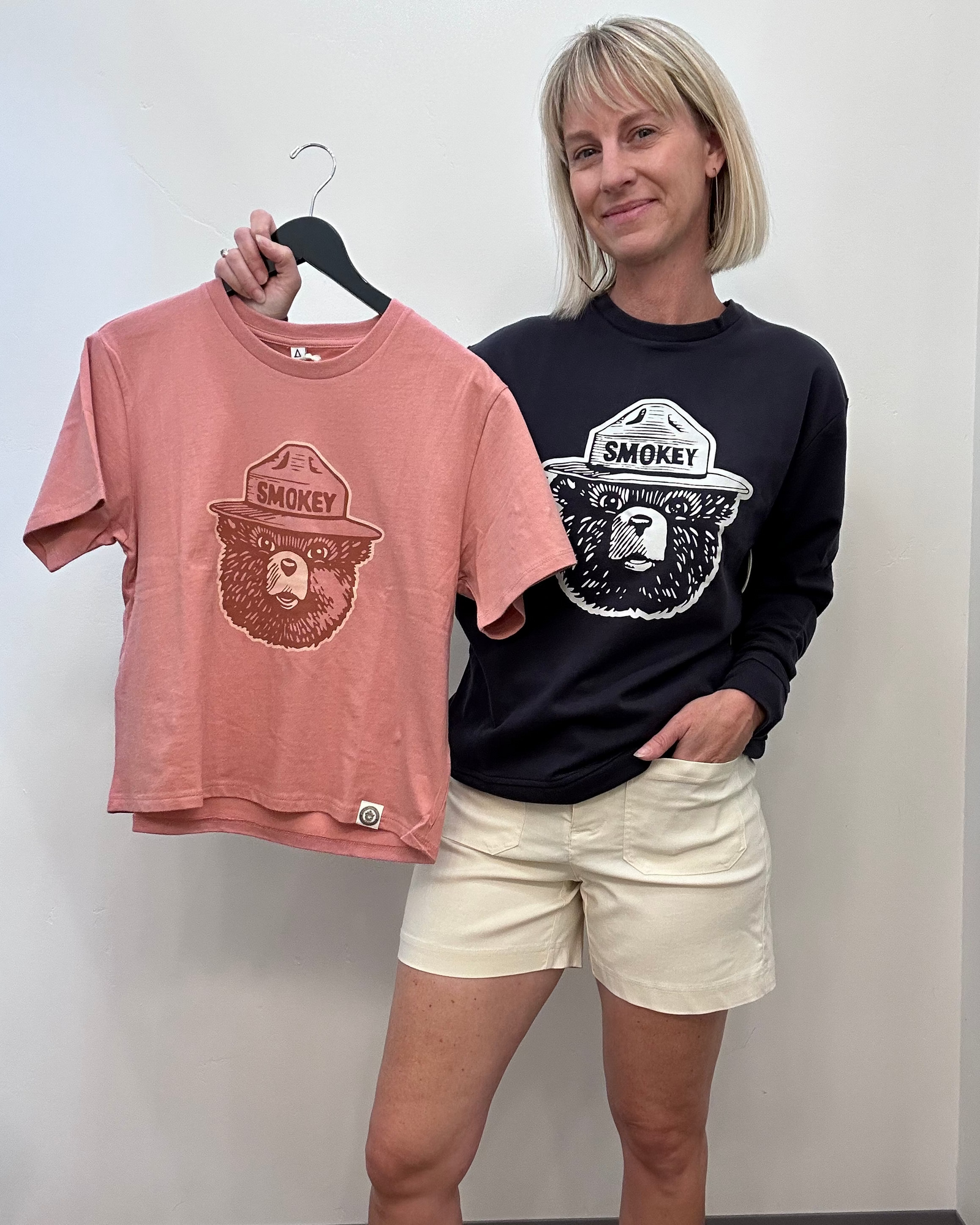 A dusty rose pink t-shirt featuring a brown graphic of a bear wearing a ranger hat with the word 'SMOKEY' on the front, styled flat-lay with dark blue jeans on a white background.
