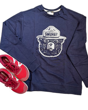 A navy blue crewneck sweatshirt featuring a white graphic of the Smokey Bear logo on the chest, styled in a flat lay with red and white running shoes on a white background.