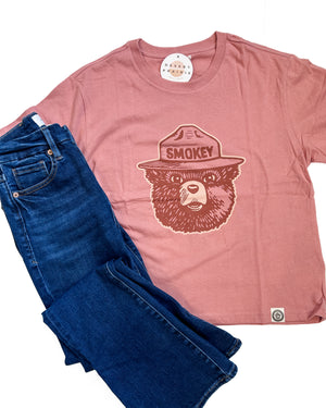 A dusty rose pink t-shirt featuring a brown graphic of a bear wearing a ranger hat with the word 'SMOKEY' on the front, styled flat-lay with dark blue jeans on a white background.