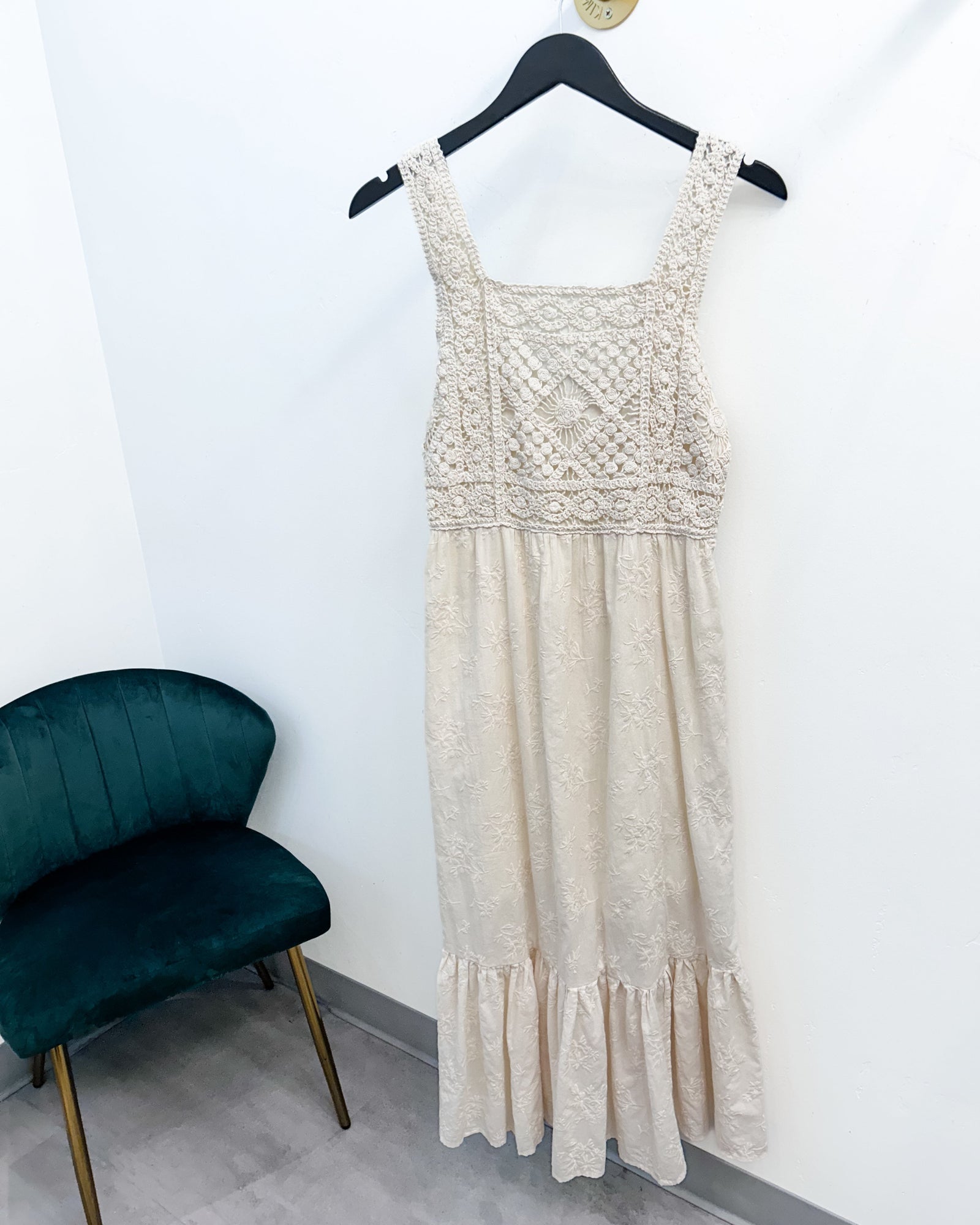 A sleeveless cream lace dress with a textured embroidered bodice and ruffled hem, hanging on a black hanger against a white wall next to a dark green velvet chair.