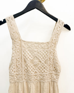 A cream-colored crochet knit dress top with a square neckline and wide straps, hanging on a black hanger against a plain white wall.