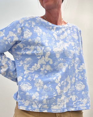 A woman wearing a light blue long-sleeve top with a white floral and sunburst pattern, paired with tan pants, shown in a mirror selfie style.