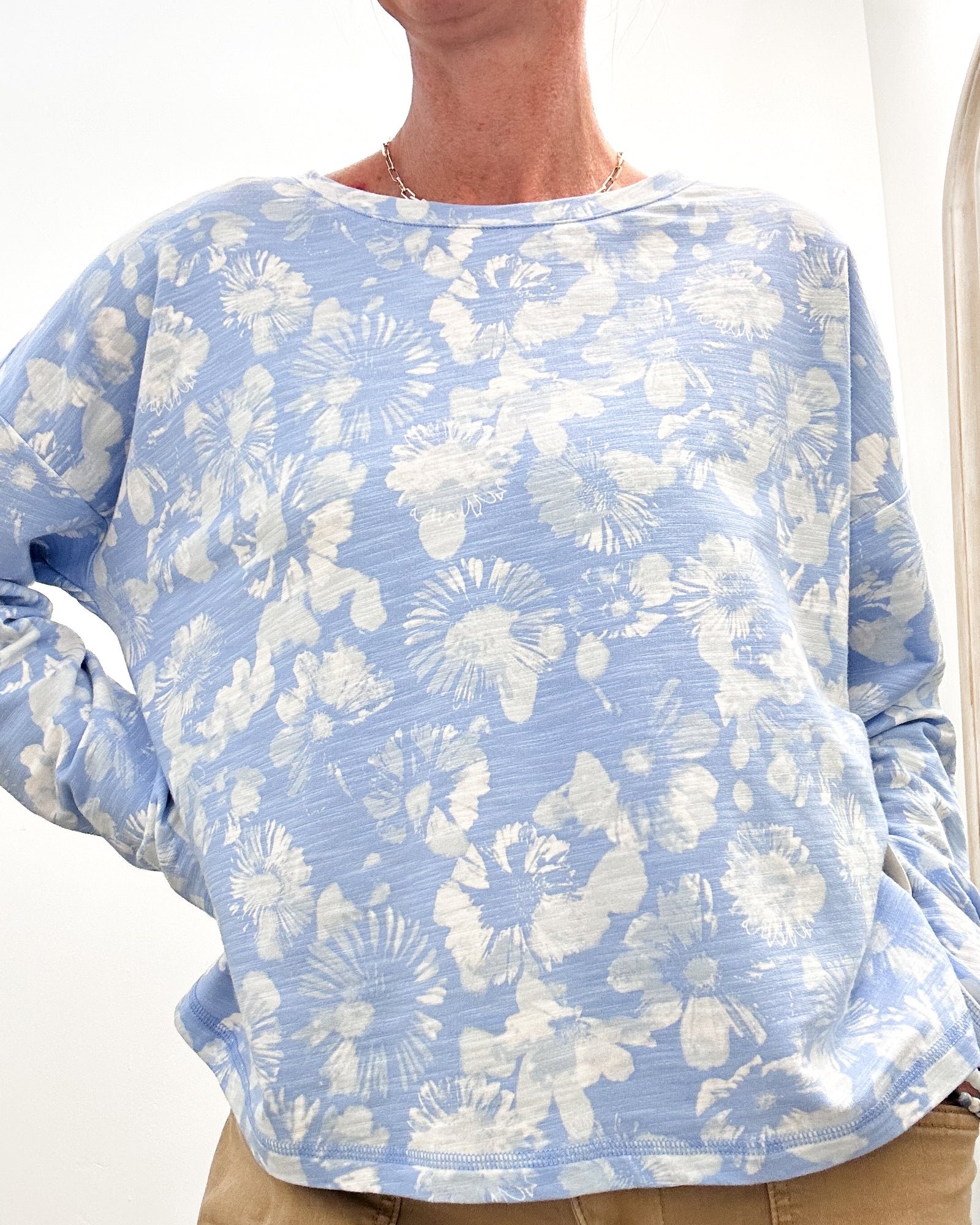A person wearing a light blue long-sleeve top with a white floral tie-dye pattern, paired with tan pants, shown against a plain white background.
