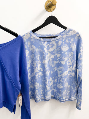 A light blue long-sleeve crop top with a white floral print hanging on a black hanger against a white wall. Another solid blue top is partially visible on the left.