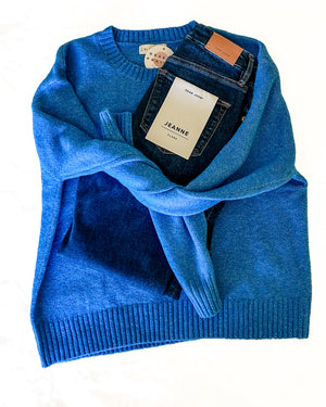 A bright blue knit sweater is laid flat on a white background, draped over a pair of dark blue jeans with visible tags including one that reads JEANNE.
