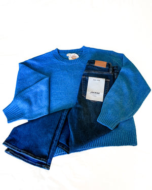 A bright blue knit crewneck sweater is laid flat on a white background, with a pair of dark blue jeans folded on top. The jeans have a white paper label that reads 'JEANNE'.