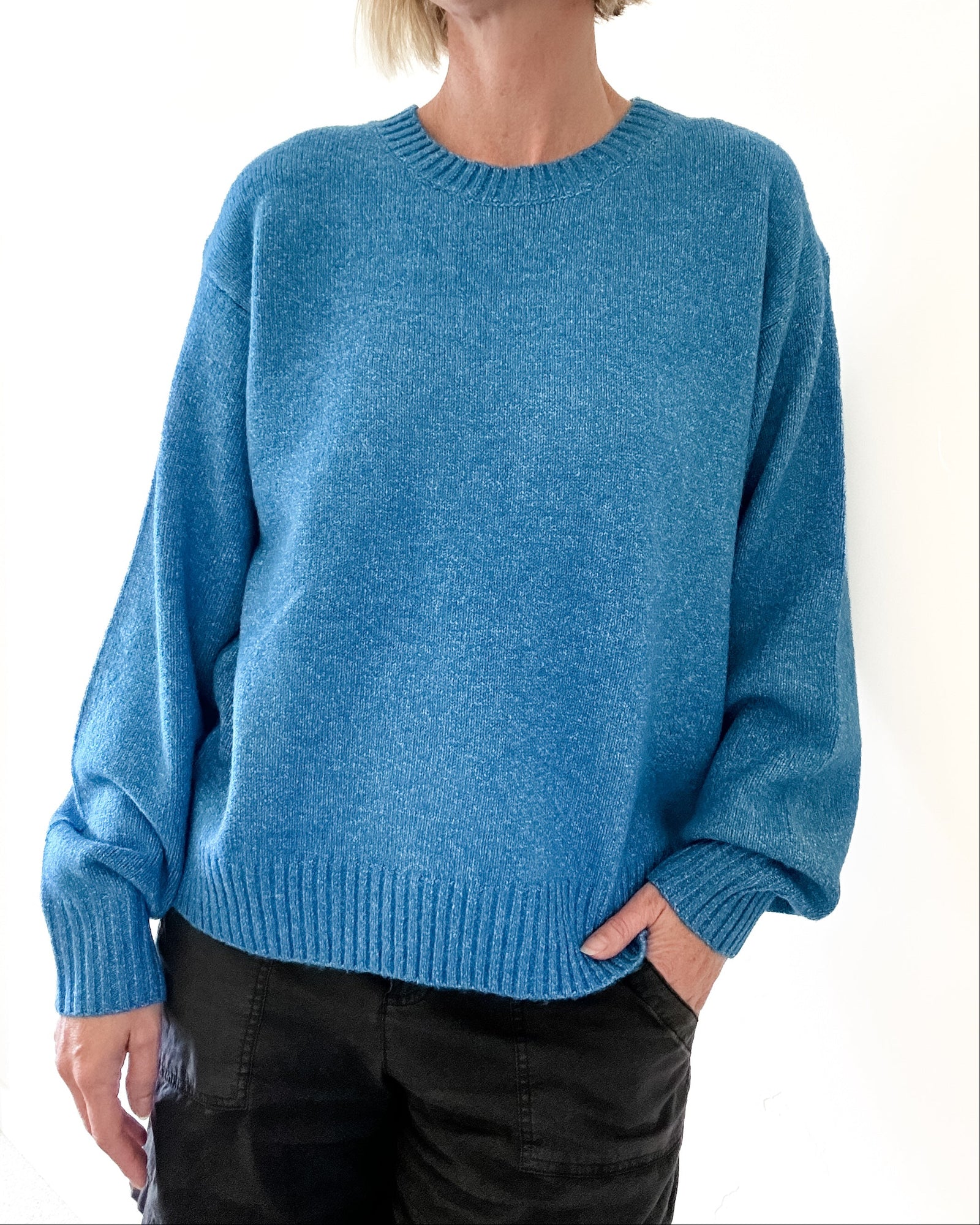 A person wearing a heathered blue crewneck knit sweater with long sleeves and ribbed cuffs and hem, paired with black pants, against a plain white background.