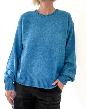 A person wearing a heathered blue crewneck knit sweater with long sleeves and ribbed cuffs and hem, paired with black pants, against a plain white background.