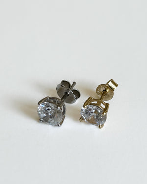 Two stud earrings on a plain light background, one in dark silver-tone metal and one in gold-tone metal, each with a clear faceted gemstone.