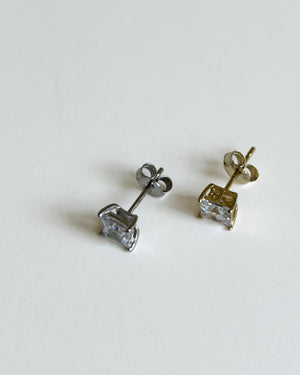 A pair of small stud earrings on a plain white background, one in silver-tone metal and one in gold-tone metal, both featuring a clear rectangular gemstone.