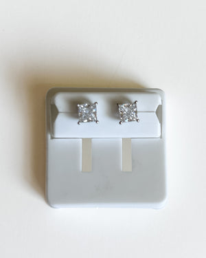 A pair of princess-cut diamond stud earrings in a silver setting, displayed on a white jewelry stand against a light beige background.
