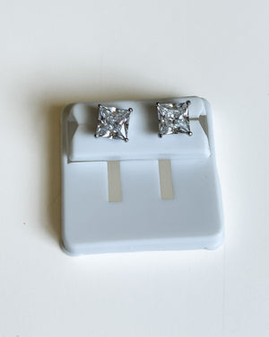 A pair of princess-cut diamond stud earrings in a silver-toned setting, displayed on a white plastic jewelry stand against a plain white background.