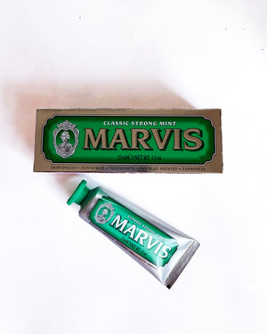 A tube of Marvis Classic Strong Mint toothpaste placed diagonally in front of its original brown and green cardboard packaging box on a plain white background.