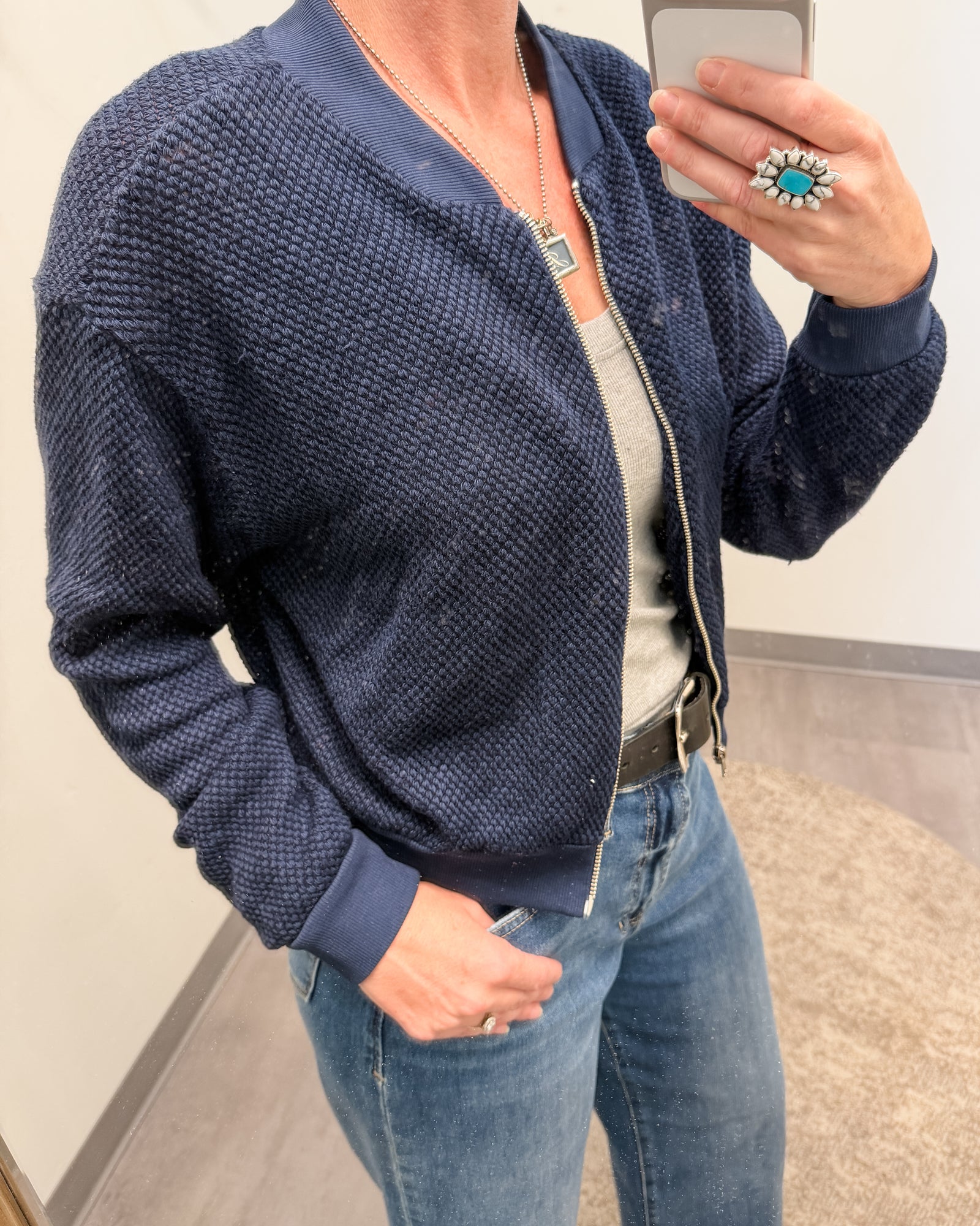 A woman takes a mirror selfie wearing a navy blue textured zip-up bomber jacket over a light gray top and blue jeans.