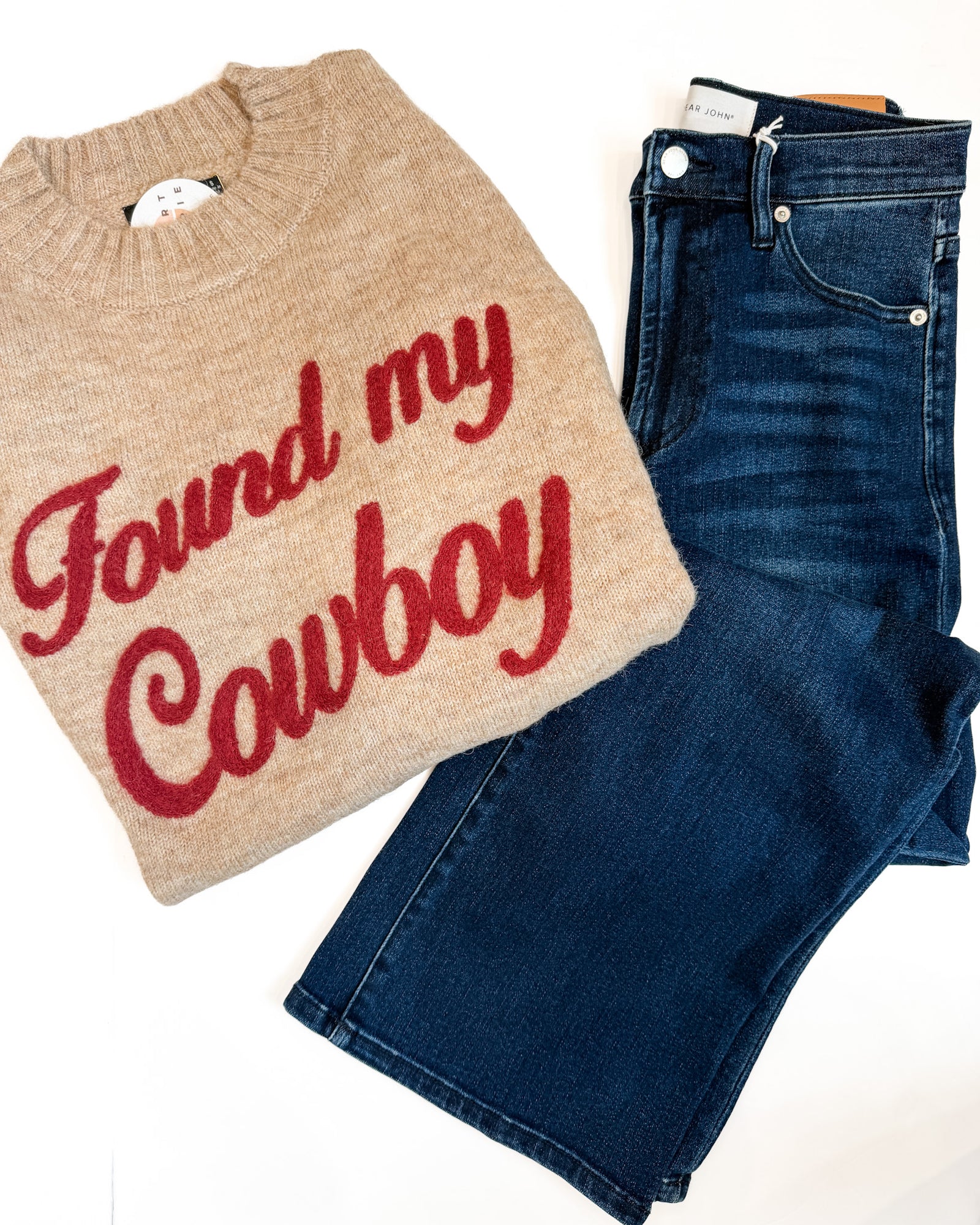 A beige knit sweater with 'Found my Cowboy' written in red script, laid flat next to a pair of dark blue jeans on a white background.
