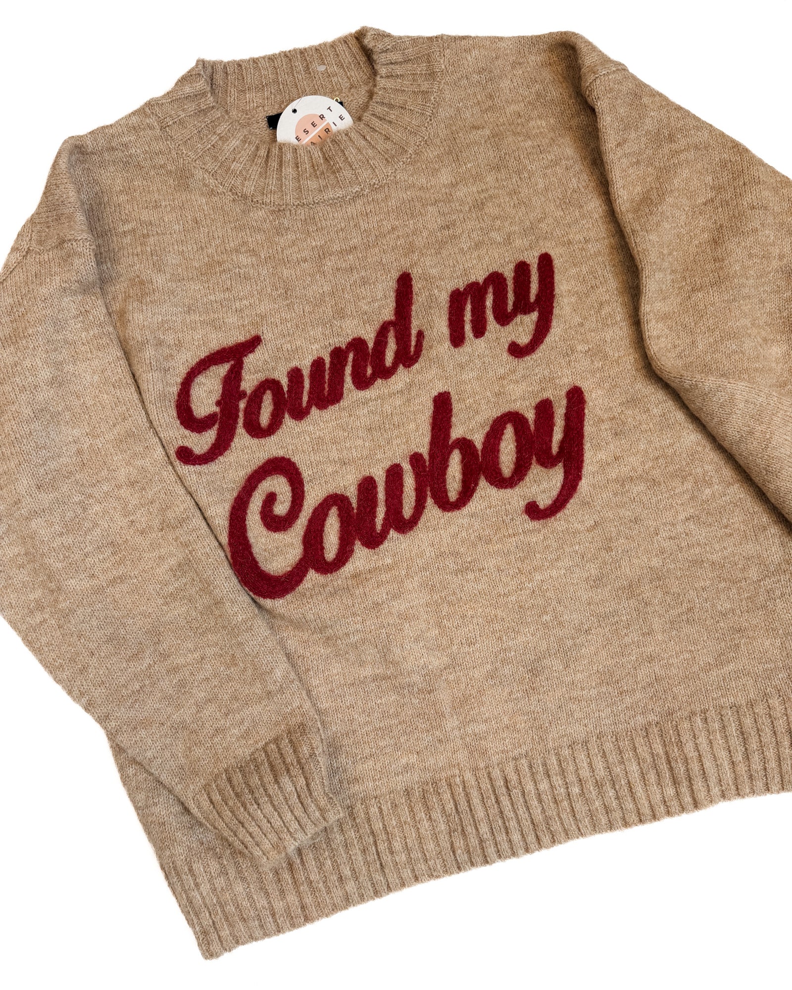 A beige knit sweater with 'Found my Cowboy' written in red script, laid flat next to a pair of dark blue jeans on a white background.