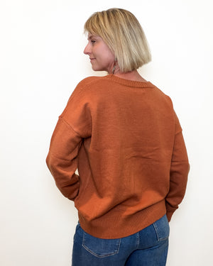 A woman with blonde hair is shown from the back wearing a rust-orange crewneck sweater and blue jeans against a plain white background.