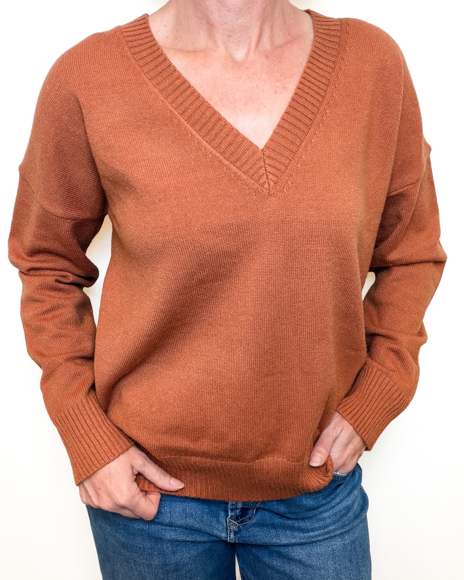 A person wearing a rust-orange ribbed V-neck sweater with long sleeves, paired with blue jeans, shown against a plain white background.